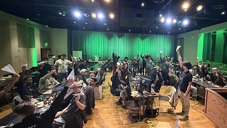 Room full of people seated at tables holding up paper airplanes