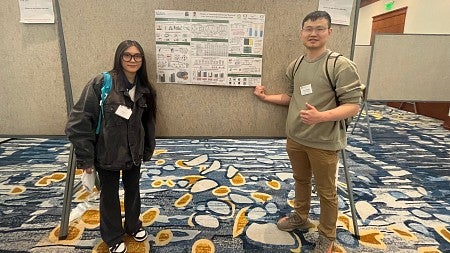 woman and man standing on either side of a large bulletin board with a poster featuring research