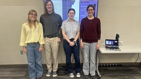 Group photo of UO students, Ayla Windley, Robert Baker, Tara Disser, and Jennifer Linnaea smiling and standing in front of a presentation screen.