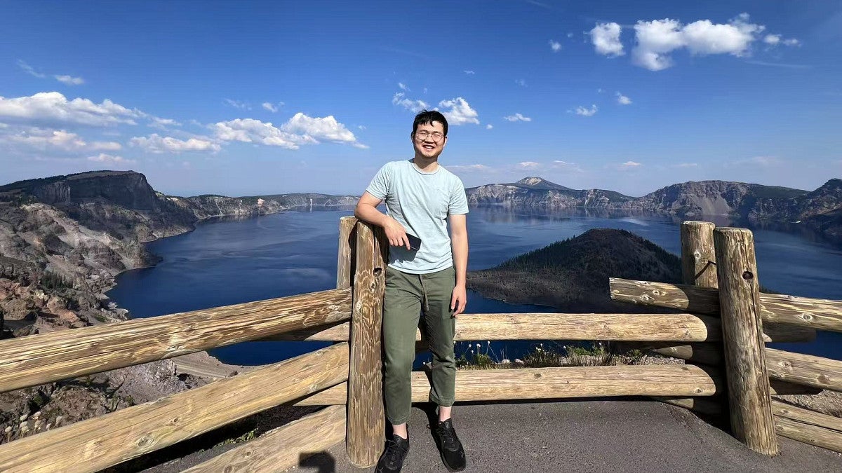 man standing against a wood fence with big blue sky and lake behind him