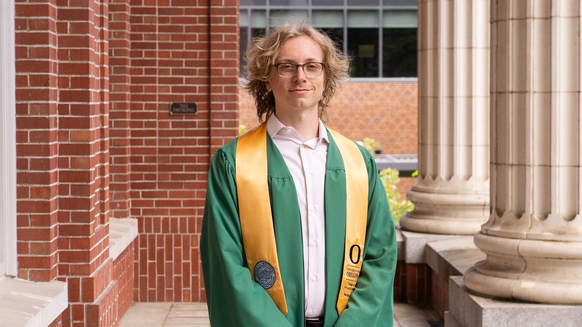 alumni noah weis standing in front of brick building in university of oregon graduation garb