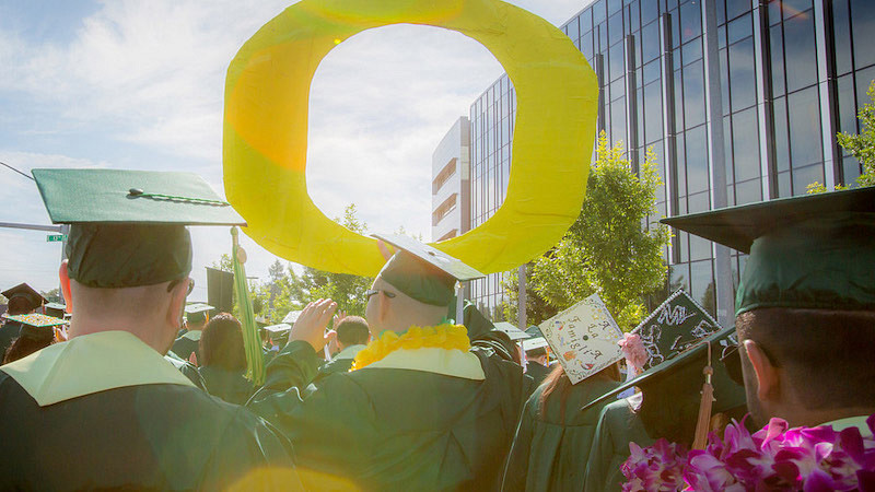 Graduates outside in green cap and gowns hold up a giant yellow O