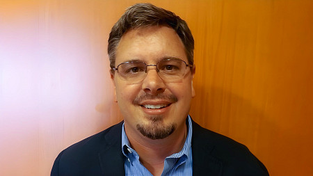 a photo of a man with facial hair and glasses smiling in front of a wood panel brown backdrop